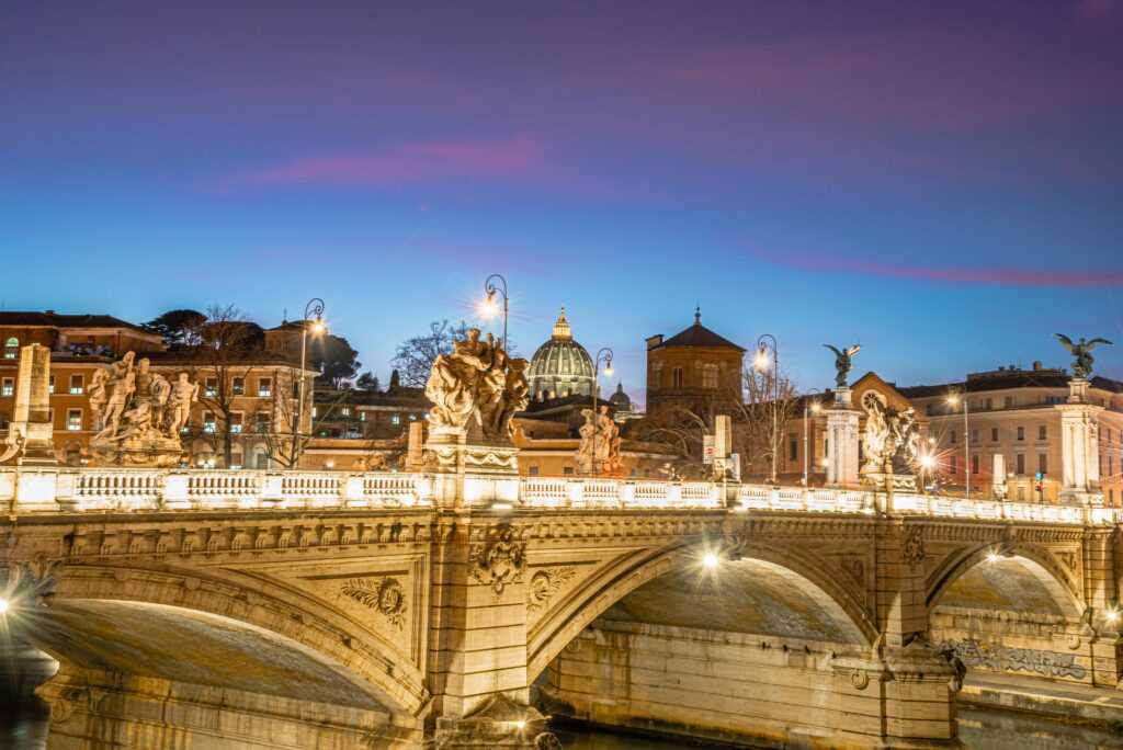 Illuminated Sant'Angelo Bridge with St. Peter's Basilica in Rome at twilight, showcasing historic architecture.